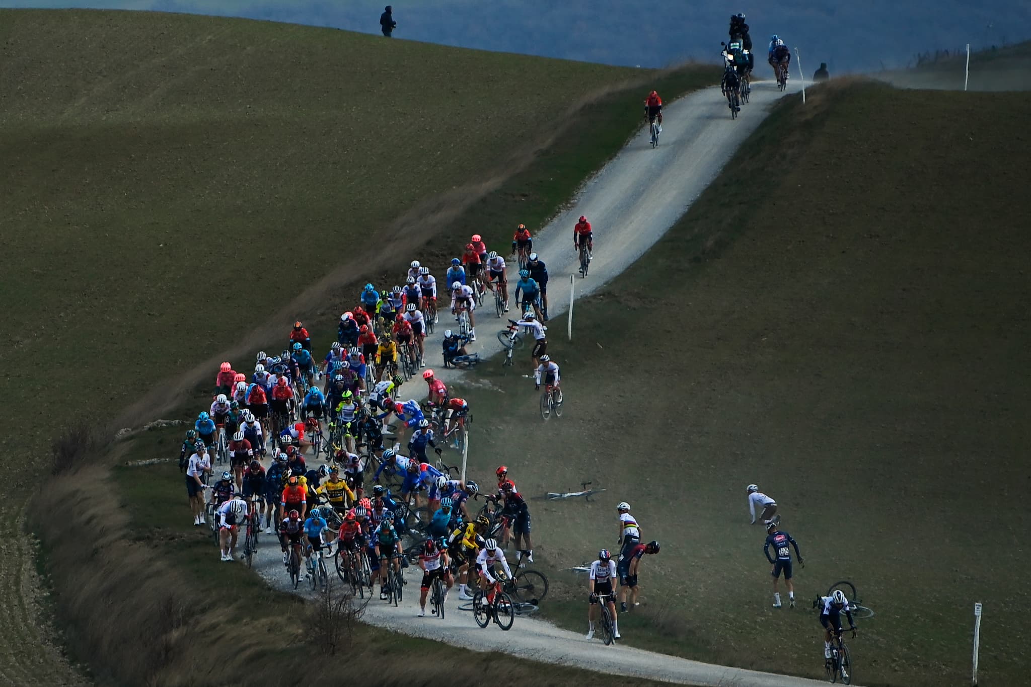 Peloton on gravel climb, crash in foreground. Strade Bianche likely.